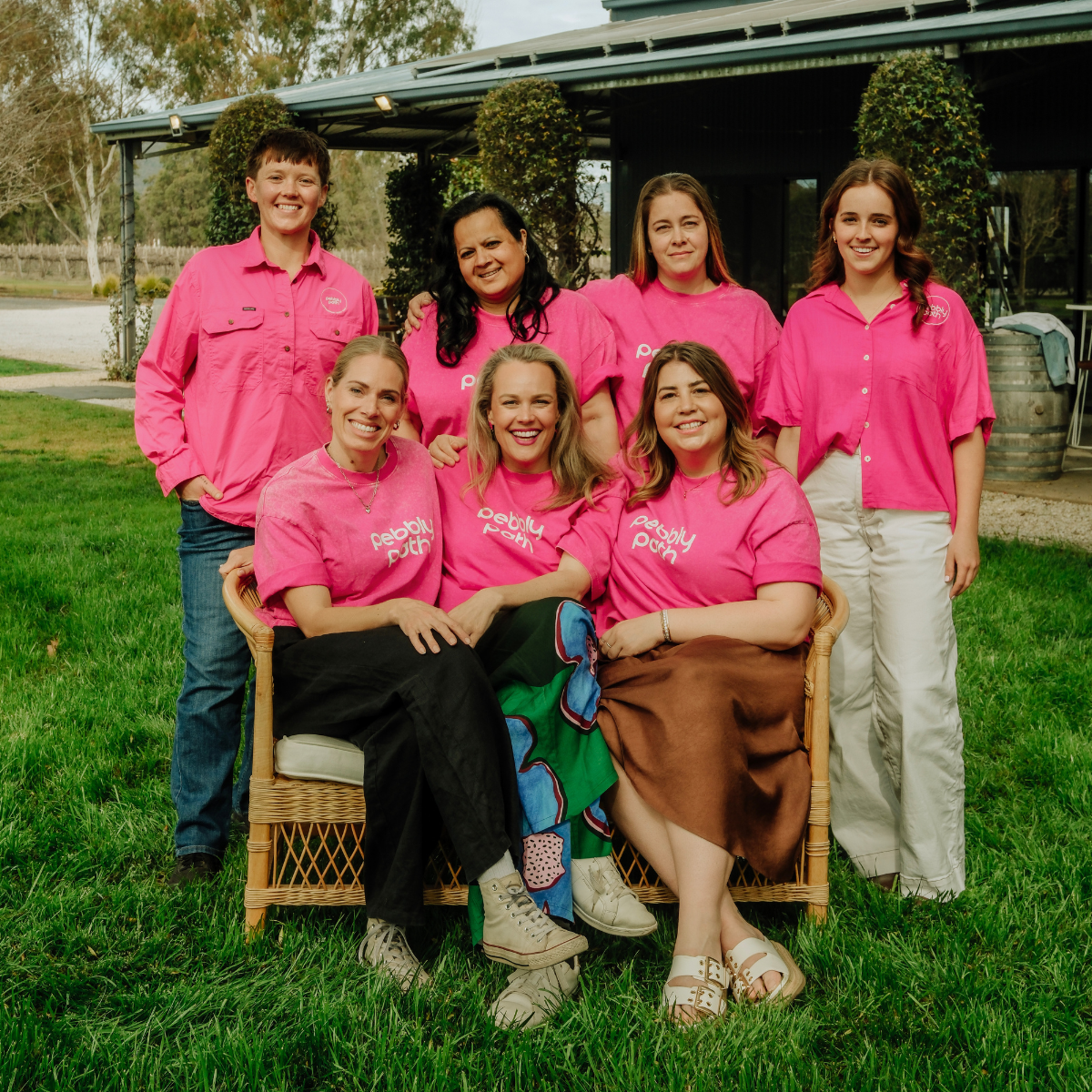 Pebbly Path team photo showing seven team members wearing bright pink branded shirts outdoors on a grassy lawn.