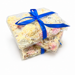 Pebbly Path White Chocolate Signature Recipe Rocky Road wrapped in clear packaging with a blue ribbon, shown from an angled view with stacked pieces, coconut flakes, and visible white chocolate coating.