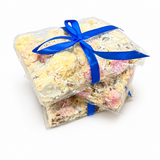 Pebbly Path White Chocolate Signature Recipe Rocky Road wrapped in clear packaging with a blue ribbon, shown from an angled view with stacked pieces, coconut flakes, and visible white chocolate coating.