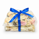 Pebbly Path White Chocolate Signature Recipe Rocky Road stacked pieces wrapped in clear packaging with a blue ribbon, shown from the front with visible coconut flakes, marshmallow, and fruit pieces on a white background.