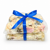 Pebbly Path White Chocolate Signature Recipe Rocky Road stacked pieces wrapped in clear packaging with a blue ribbon, shown from the front with visible coconut flakes, marshmallow, and fruit pieces on a white background.