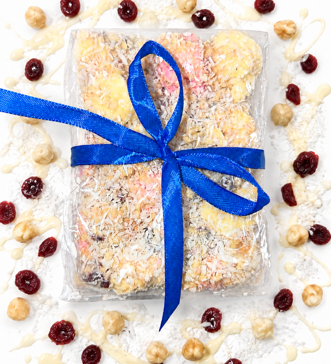 Pebbly Path White Chocolate Signature Recipe Rocky Road wrapped in clear packaging with a blue ribbon, topped with coconut flakes, surrounded by dried cranberries, macadamias, and white chocolate drizzle on a white background.