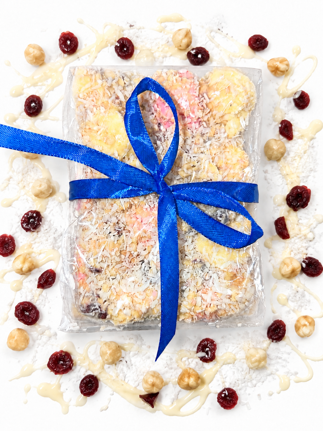 Pebbly Path White Chocolate Signature Recipe Rocky Road wrapped in clear packaging with a blue ribbon, topped with coconut flakes, surrounded by dried cranberries, macadamias, and white chocolate drizzle on a white background.