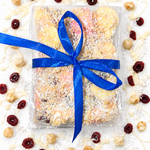Pebbly Path White Chocolate Signature Recipe Rocky Road wrapped in clear packaging with a blue ribbon, topped with coconut flakes, surrounded by dried cranberries, macadamias, and white chocolate drizzle on a white background.