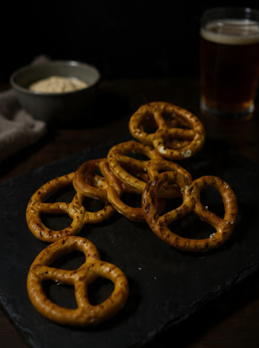 Pretzels arranged on a dark serving board with sea salt flakes and a warm drink in the background for a savoury snack display