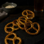 Pretzels arranged on a dark serving board with sea salt flakes and a warm drink in the background for a savoury snack display