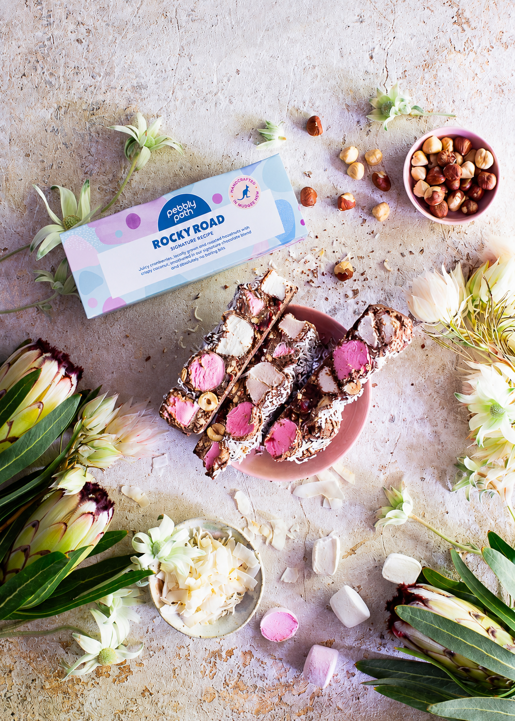 Pebbly Path Signature Recipe Rocky Road 100g bars styled with pink marshmallows, hazelnuts, coconut flakes, and branded packaging surrounded by native flowers and confectionery ingredients.