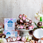 Pebbly Path Signature Recipe Rocky Road stacked on a cake stand with marshmallows, hazelnuts, coconut, fresh flowers, and branded packaging in a styled product display.