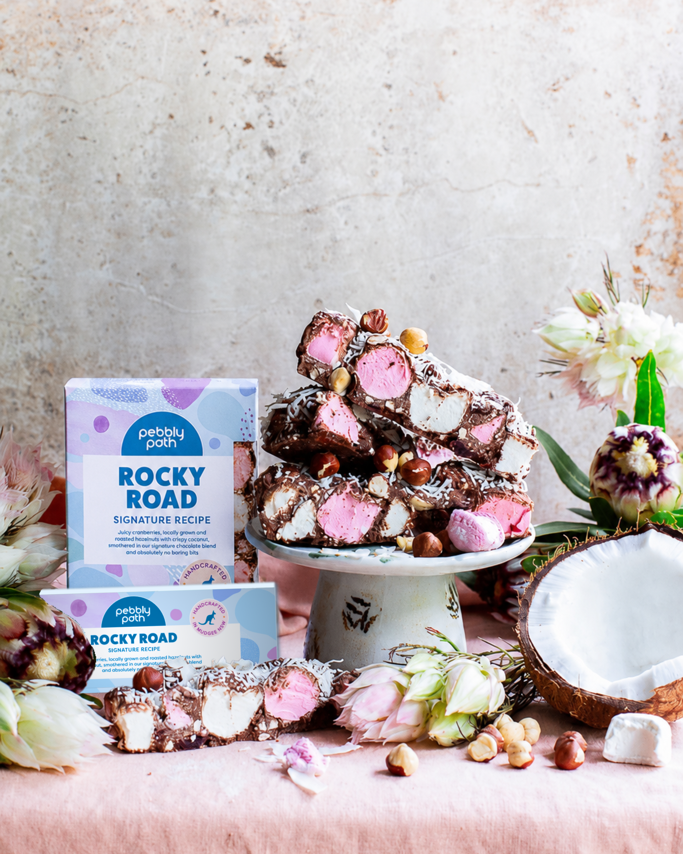 Pebbly Path Signature Recipe Rocky Road stacked on a cake stand with marshmallows, hazelnuts, coconut, fresh flowers, and branded packaging in a styled product display.