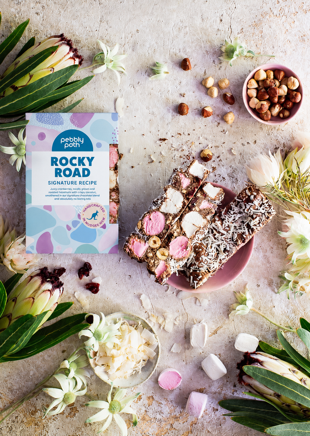 Pebbly Path Signature Recipe Rocky Road bars styled with marshmallows, hazelnuts, coconut flakes, dried cranberries, and branded packaging surrounded by native flowers.