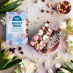 Pebbly Path Signature Recipe Rocky Road bars styled with marshmallows, hazelnuts, coconut flakes, dried cranberries, and branded packaging surrounded by native flowers.