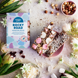 Pebbly Path Signature Recipe Rocky Road bars styled with marshmallows, hazelnuts, coconut flakes, dried cranberries, and branded packaging surrounded by native flowers.