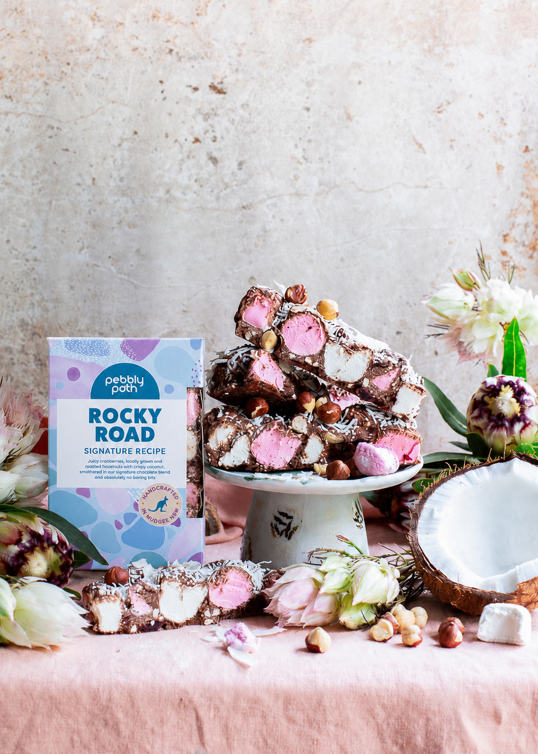 Pebbly Path Signature Recipe Rocky Road displayed with chunky marshmallows, roasted hazelnuts, coconut flakes, fresh flowers, and branded gift packaging on a styled dessert table.