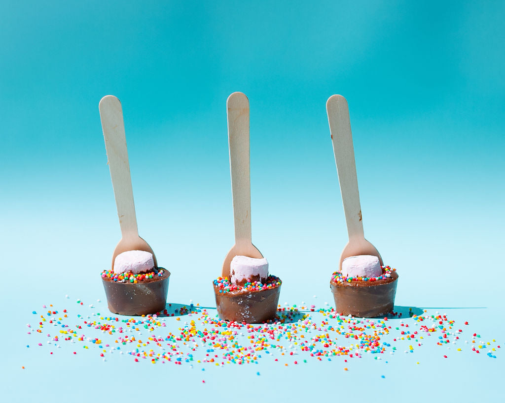 Pebbly Path Hot Chocolate Pops with marshmallows and colourful sprinkles displayed on a pastel blue background with scattered rainbow sprinkles