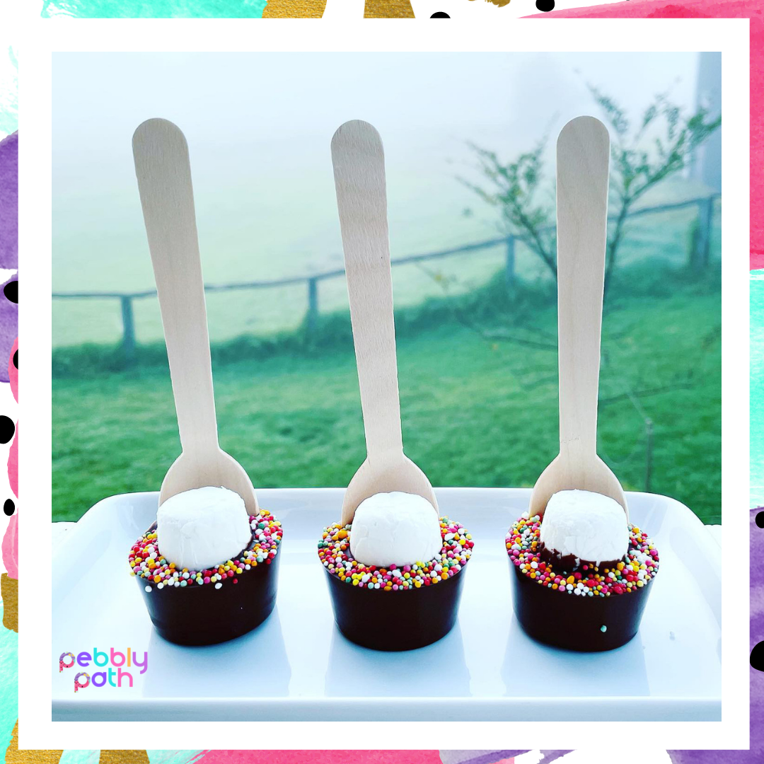 Pebbly Path Hot Chocolate Pops with marshmallows and colourful sprinkles displayed as a trio on a white tray with an outdoor countryside backdrop